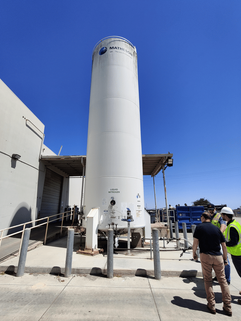 Men in protective gear meet near a tall cylinder tank at an industrial site